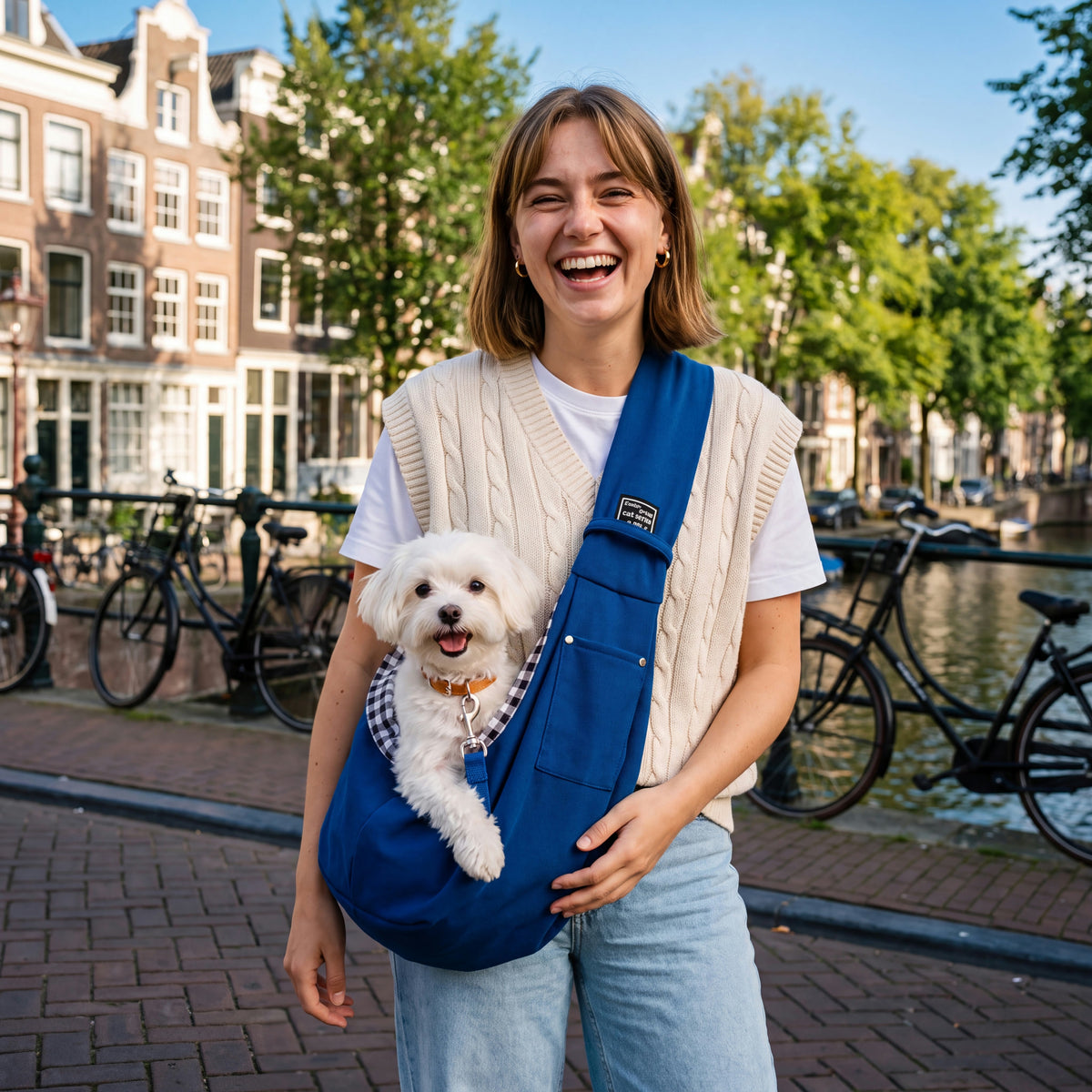 Jonge vrouw op een grachtbrug met de blauwe CuddleCarry™ schoudertas; een witte maltezer kijkt blij over de rand.