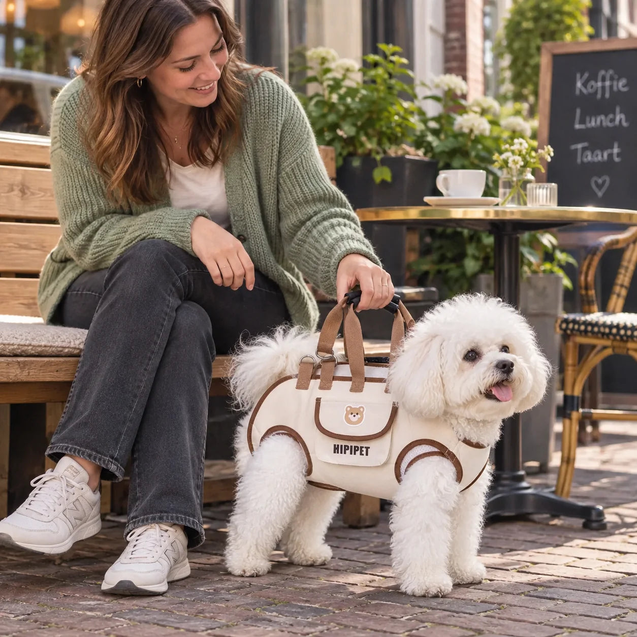 Baasje met de crèmekleurige draagtas met pootopeningen bij een café; de hond staat ernaast op de grond.