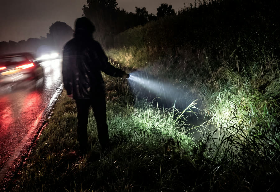 Silhouet met een zaklamp zoekt in de berm langs een natte weg, koplampen komen voorbij.